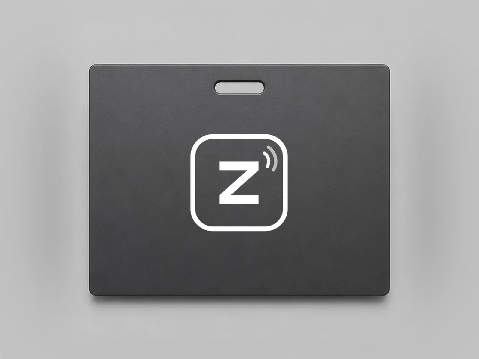 Matte graphite NFC name badge with a lanyard slot at the top and the white Zovo emblem centered on its face, on a light grey studio backdrop